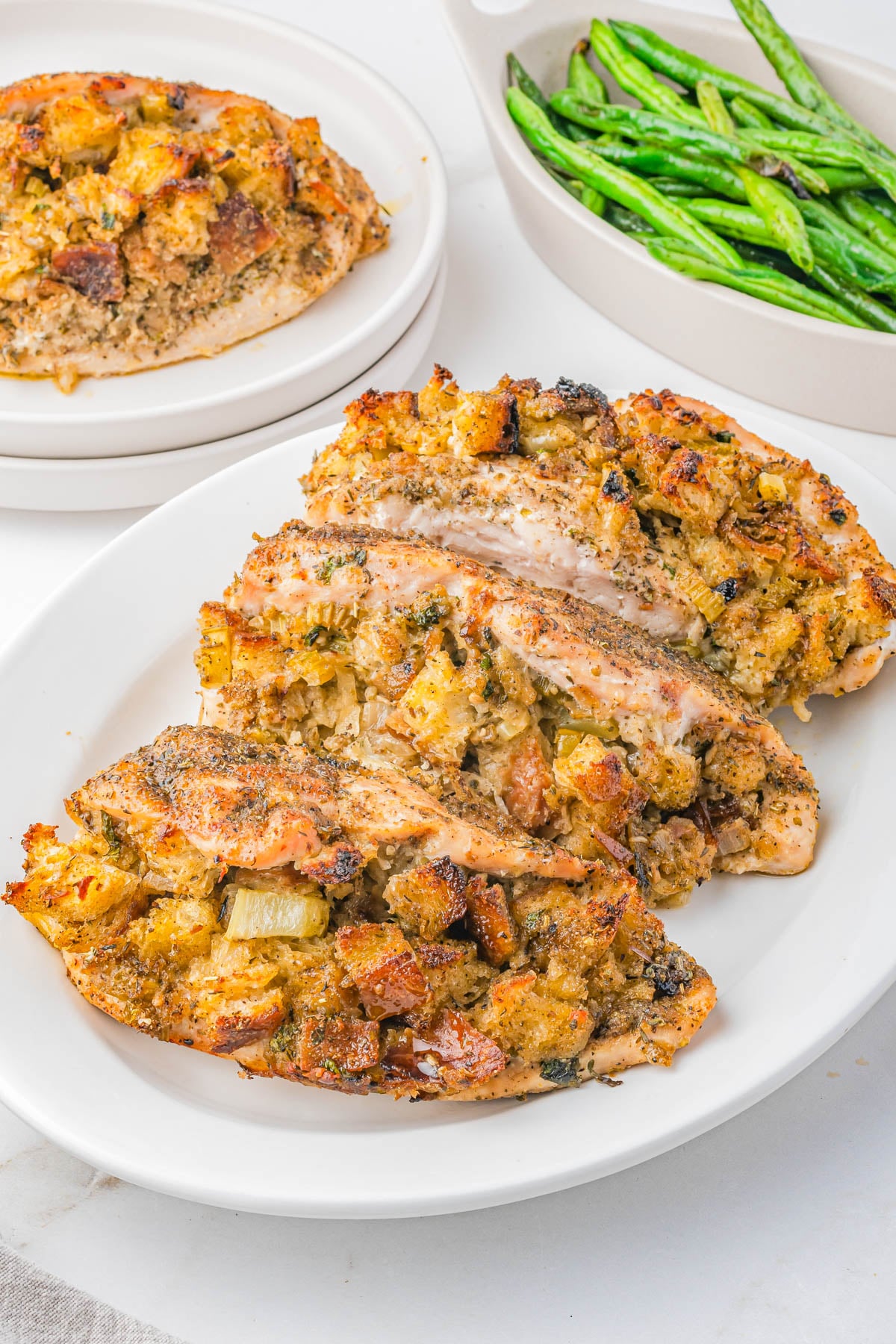 Three stuffed chicken breasts with bread stuffing are arranged on a white plate, with a side dish of green beans and an additional serving on a separate plate in the background.