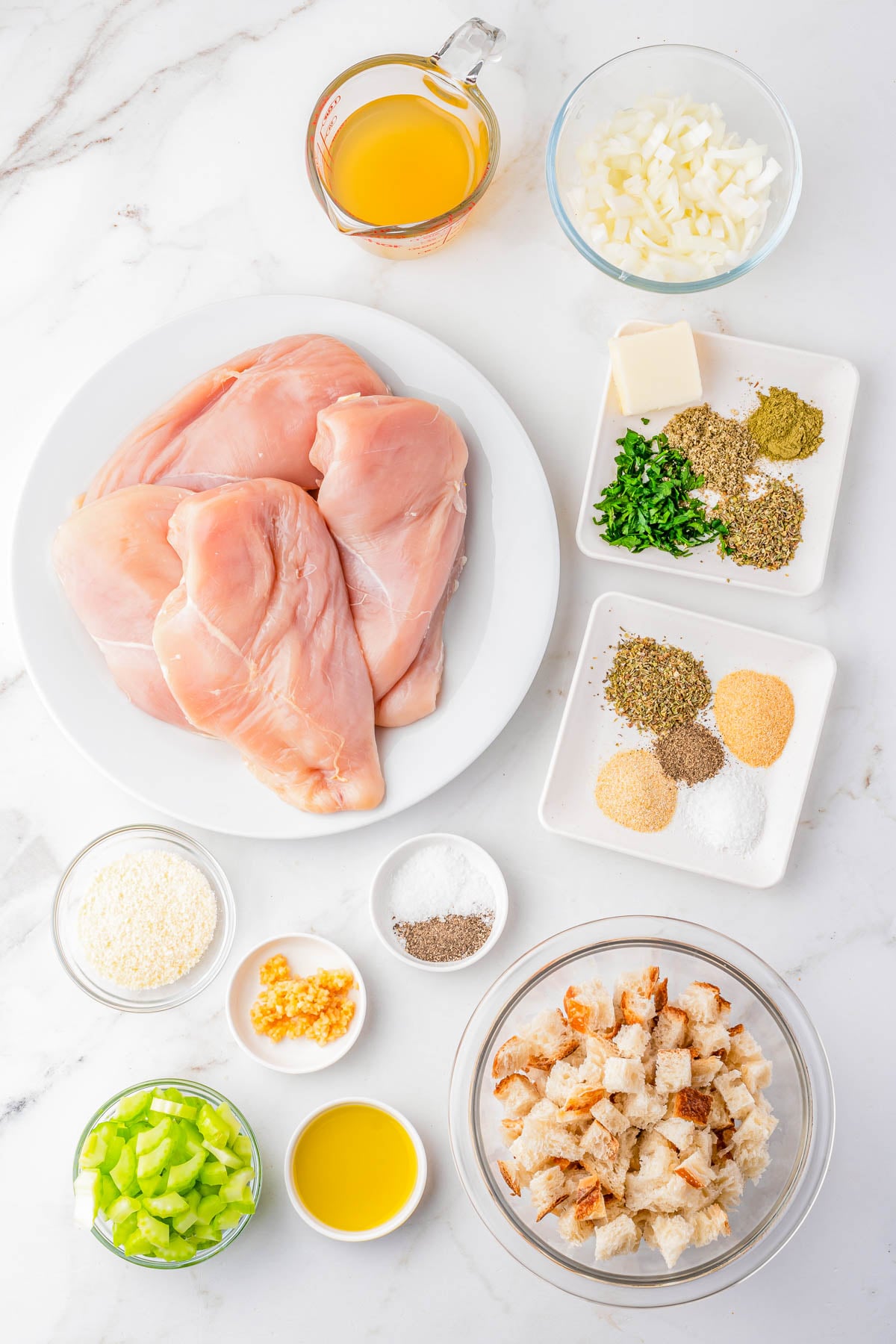 Raw chicken breasts, chopped onions, celery, herbs, spices, garlic, butter, oil, chicken broth, breadcrumbs, grated cheese, salt, and pepper arranged in bowls and plates on a marble surface.