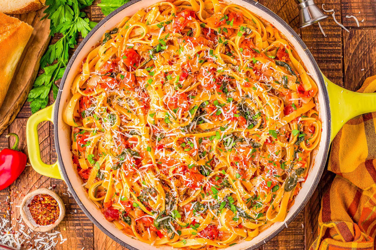 Large pan of fettuccine pasta mixed with tomato sauce, fresh herbs, and grated cheese, surrounded by bread, parsley, chili flakes, and a yellow napkin on a wooden table.
