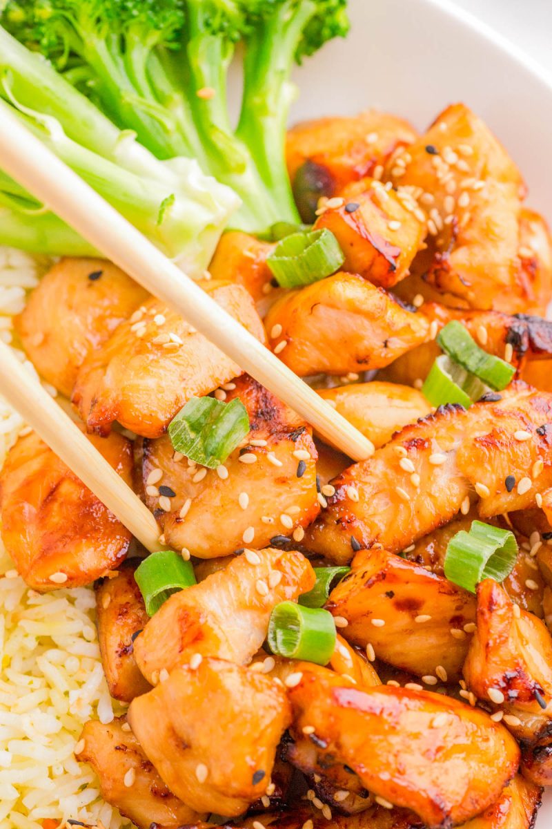 Chopsticks holding grilled chicken pieces topped with sesame seeds and green onions, served with rice and steamed broccoli on a white plate.