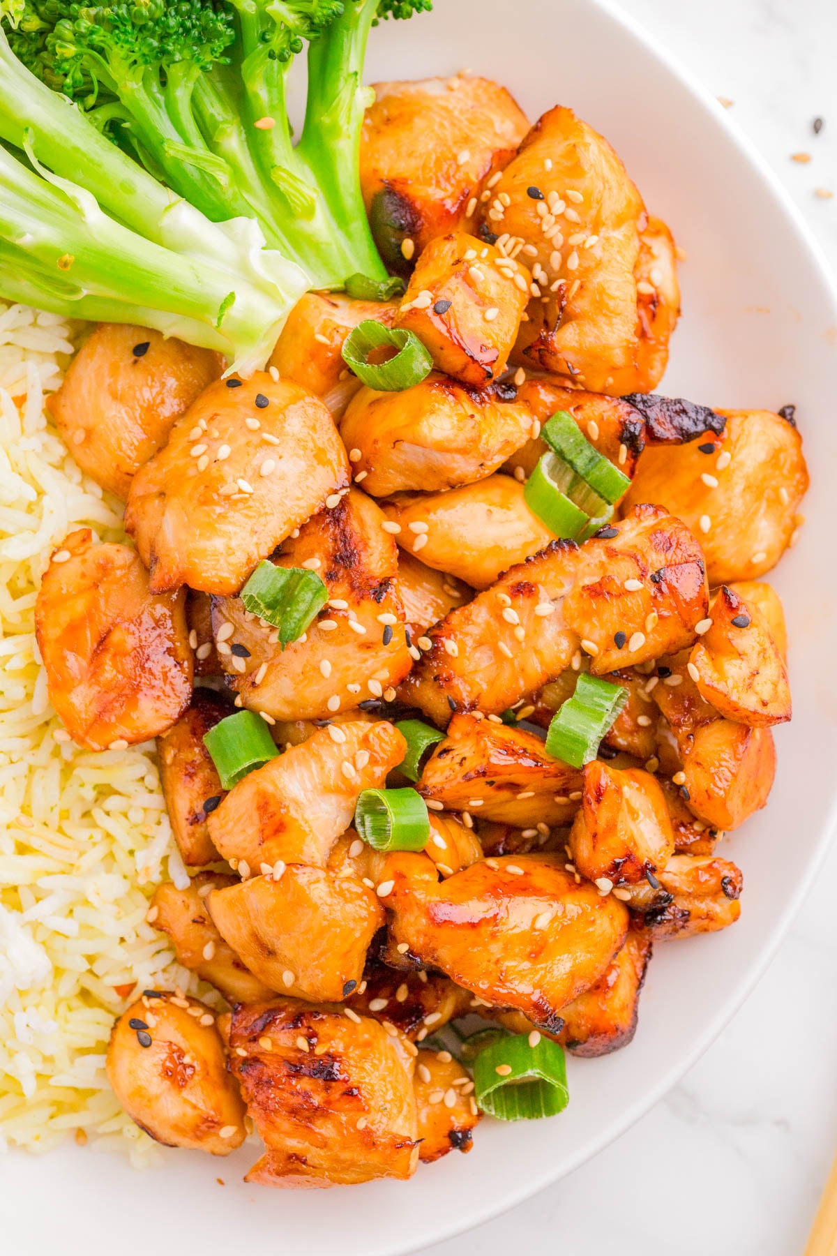 A plate of grilled chicken pieces topped with sesame seeds and chopped green onions, served with steamed broccoli and yellow rice.