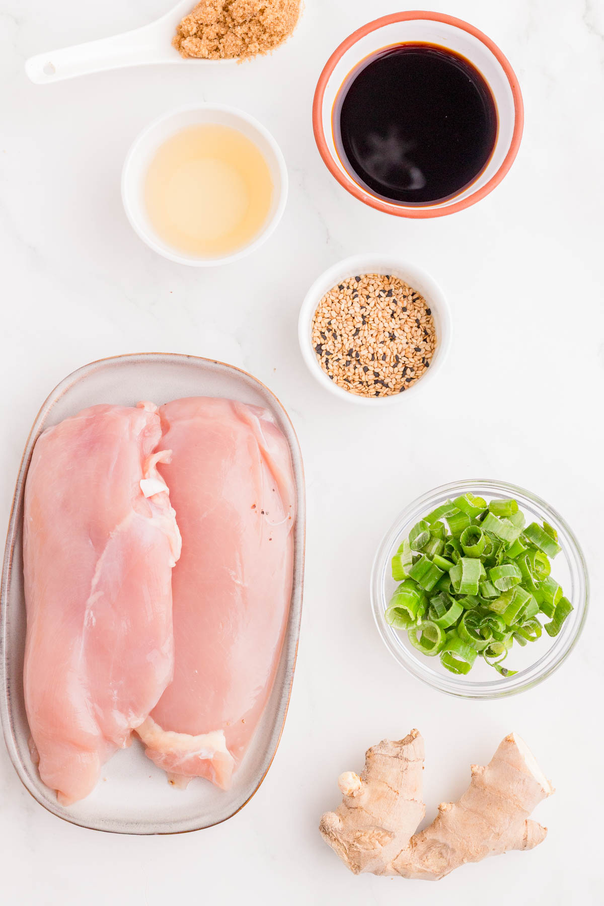 Raw chicken breasts on a plate with bowls of sliced green onions, brown sugar, soy sauce, sesame seeds, rice vinegar, and a piece of fresh ginger on a white surface.