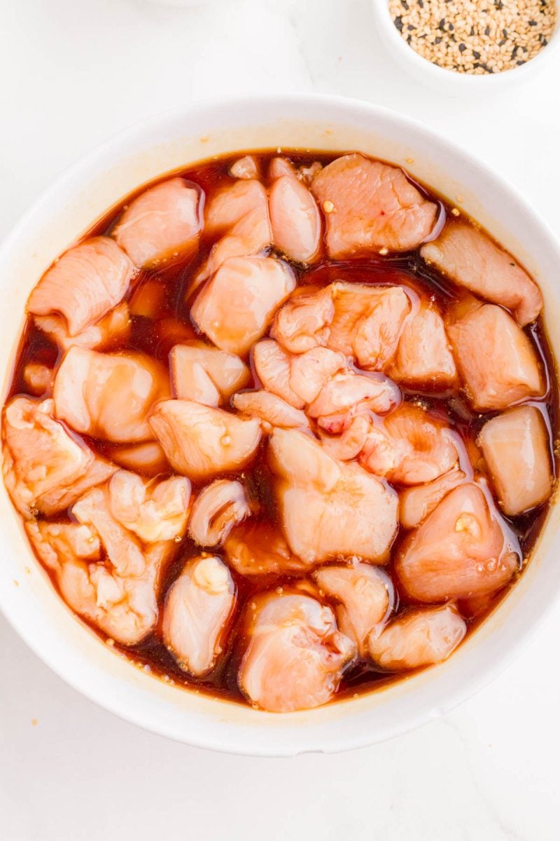 Chunks of raw chicken breast marinating in a brown sauce inside a white bowl, viewed from above.