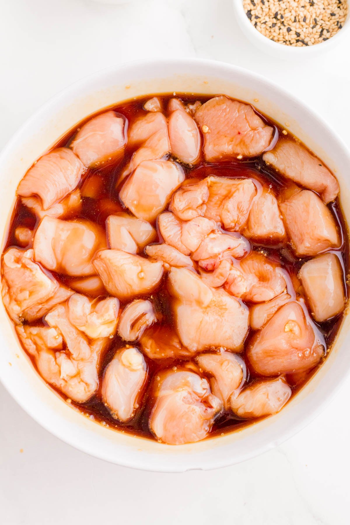 Chunks of raw chicken breast marinating in a brown sauce inside a white bowl, viewed from above.