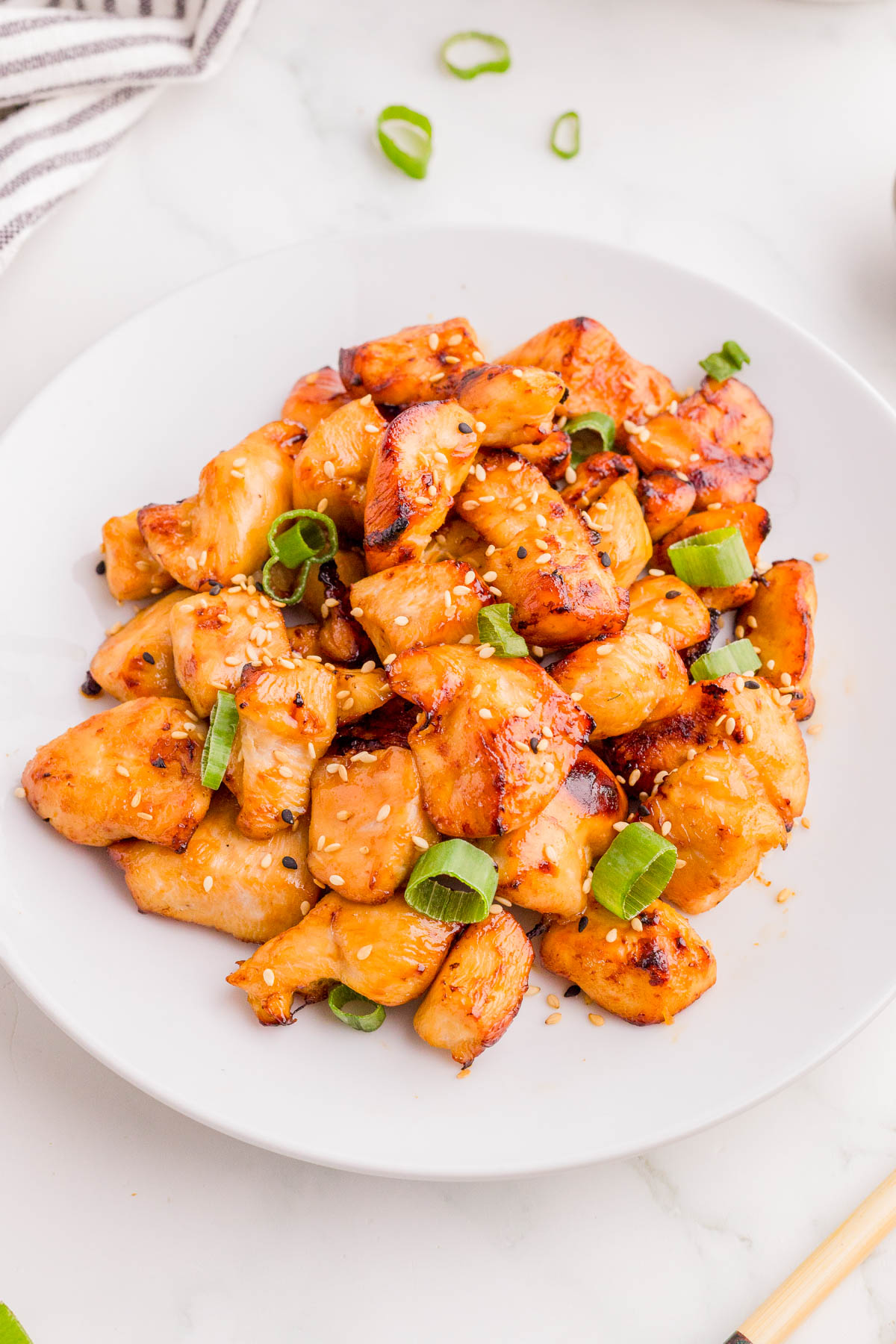 A white plate filled with cooked teriyaki chicken pieces, garnished with sliced green onions and sesame seeds.