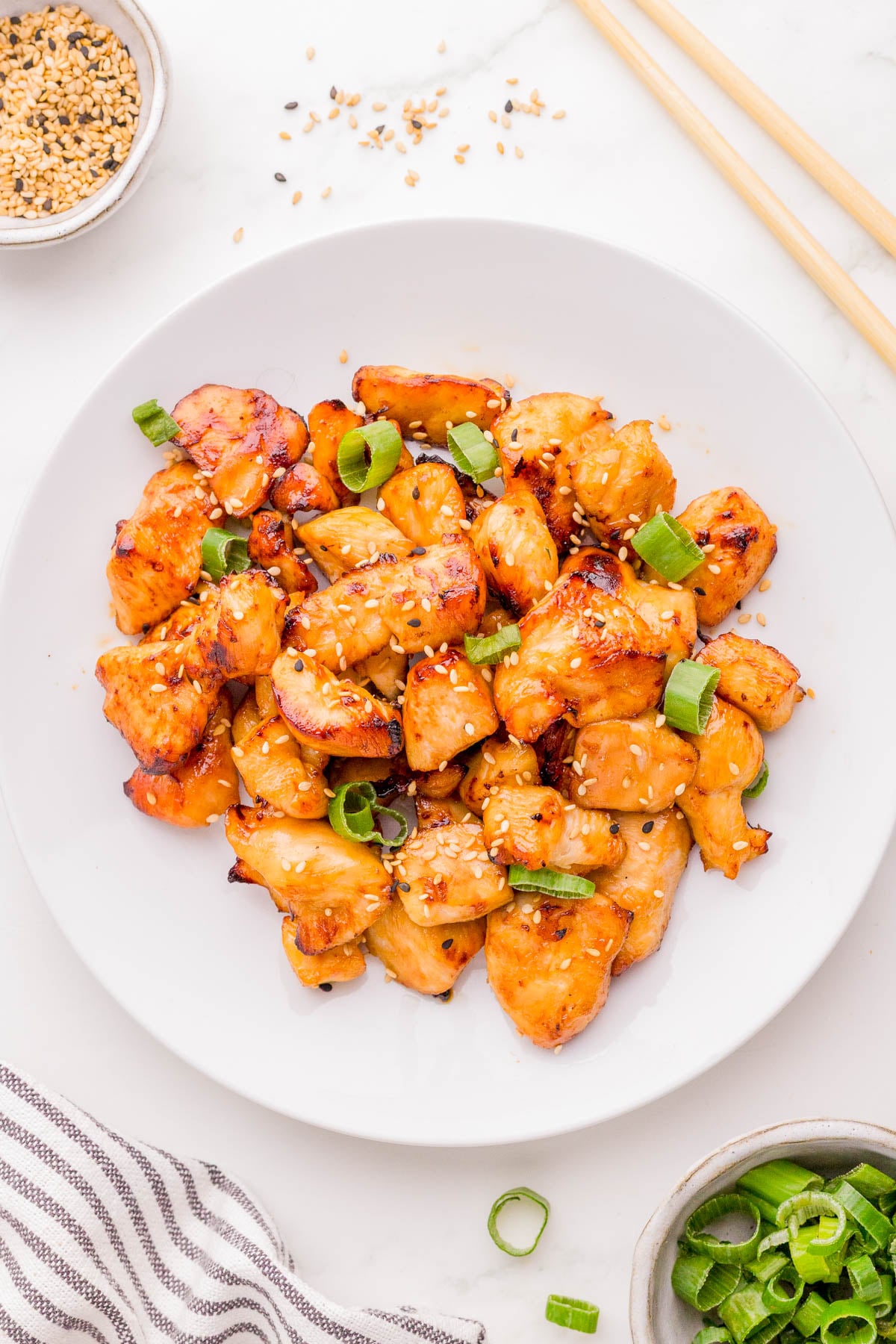 A white plate with cooked pieces of glazed chicken, garnished with sesame seeds and chopped green onions, set on a white surface with chopsticks and a small bowl of sesame seeds nearby.