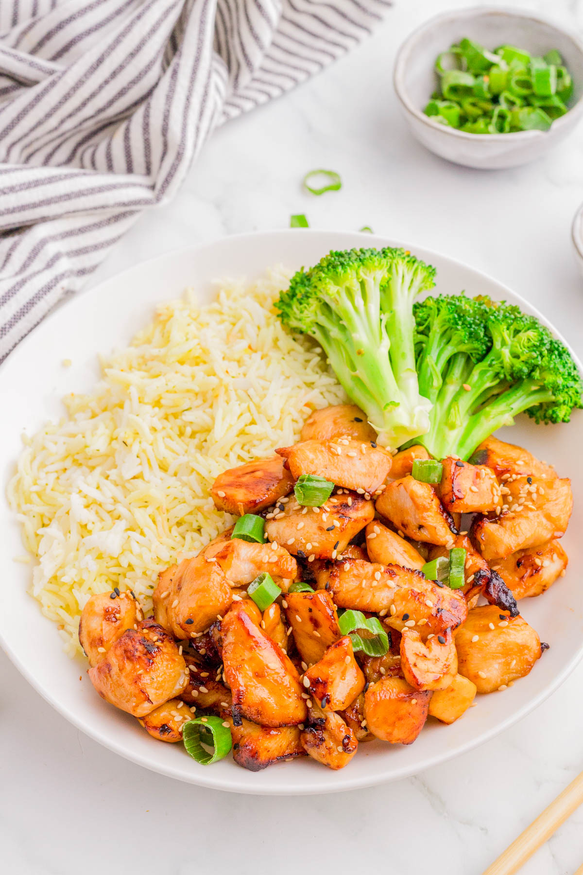 A plate with grilled chicken pieces topped with green onions, served with steamed broccoli and white rice on the side.