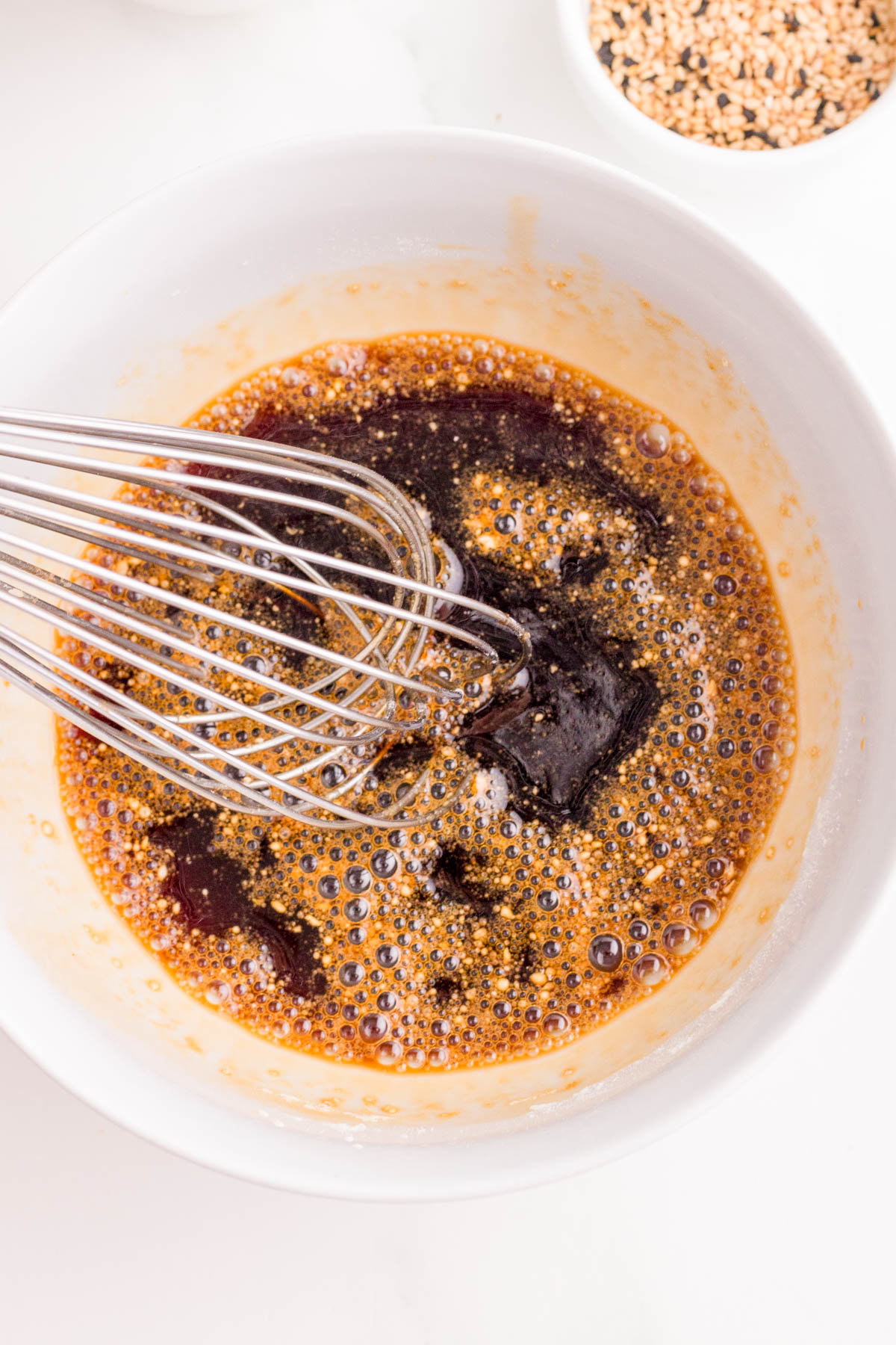 A metal whisk mixes a brown, bubbly liquid mixture in a white bowl, with a small bowl of sesame seeds visible in the background.