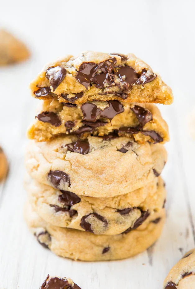 A stack of five chocolate chip cookies on a white surface, with the top cookie broken in half to show melted chocolate chips inside.