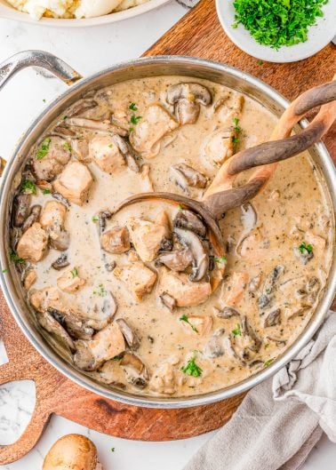 A skillet filled with creamy mushroom and chicken stew, garnished with chopped parsley, with a wooden spoon and ladle resting in the pan.