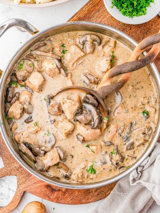 A skillet filled with creamy mushroom and chicken stew, garnished with chopped parsley, with a wooden spoon and ladle resting in the pan.