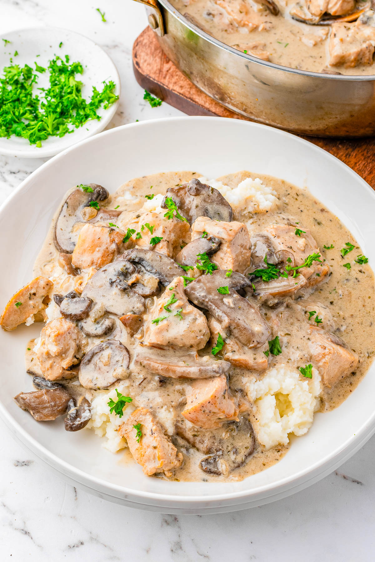 A plate of mashed potatoes topped with creamy chicken and mushroom sauce, garnished with chopped parsley. A pan of the same dish and a small plate of parsley are in the background.