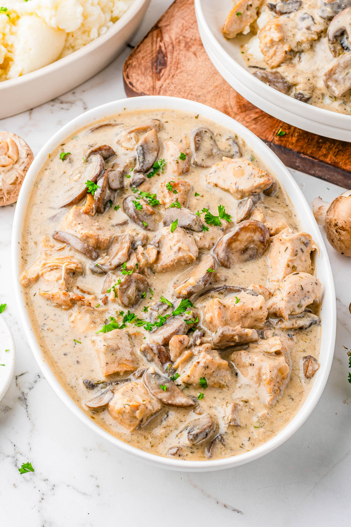 A white oval dish filled with chicken and mushrooms in a creamy sauce, garnished with chopped parsley, sits on a marble countertop.