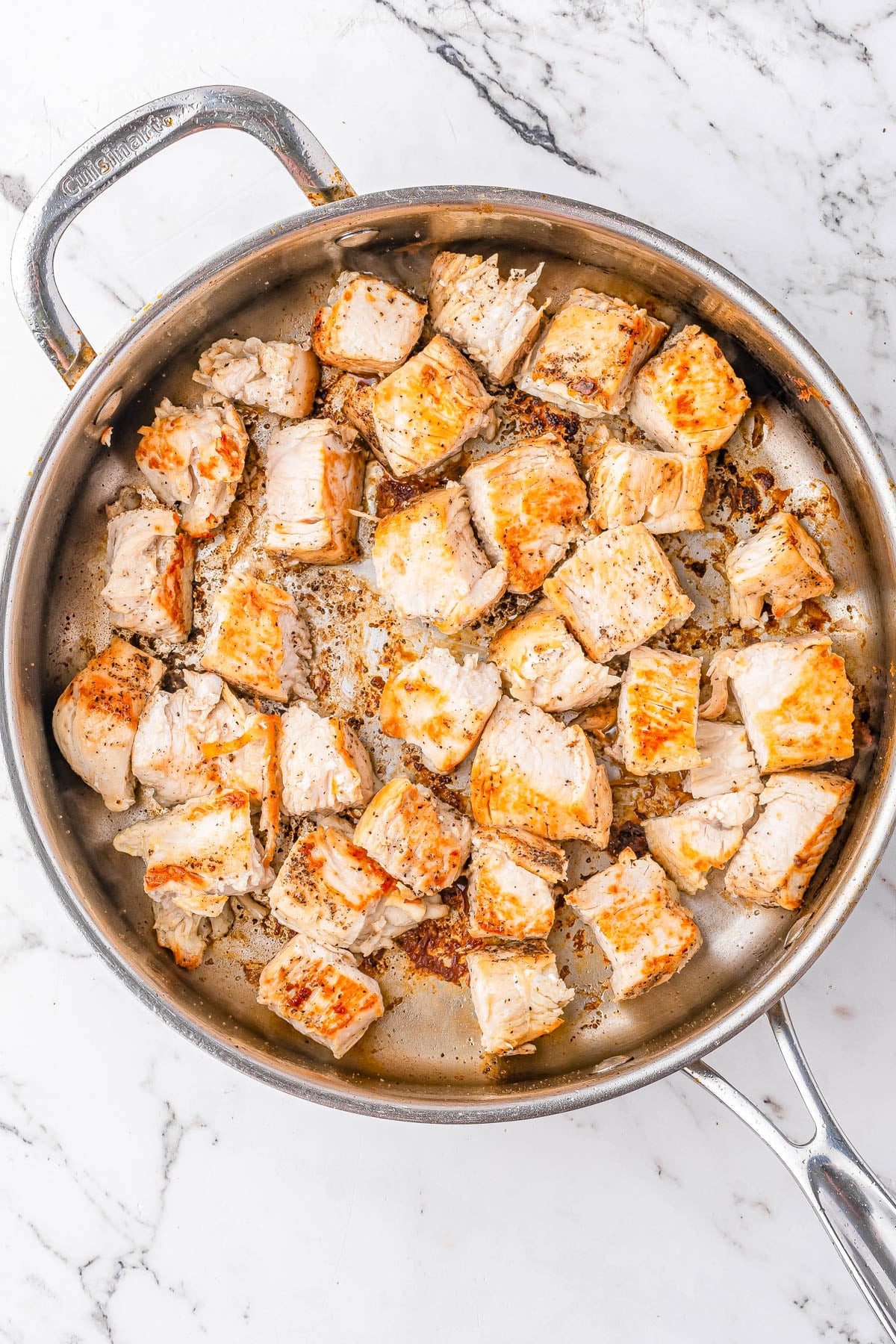 Chunks of browned chicken cooking in a stainless steel skillet on a marble countertop.
