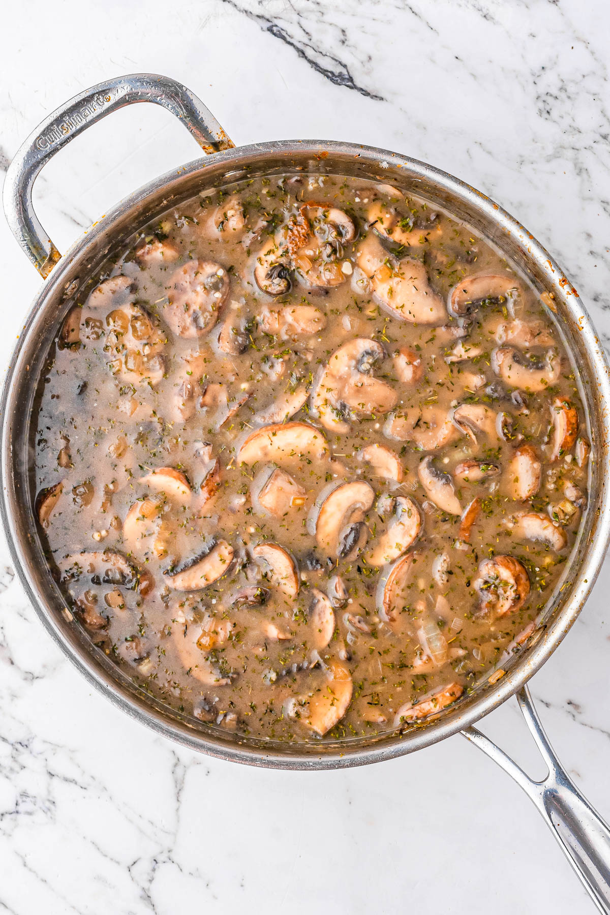 A stainless steel pan filled with sliced mushrooms in a brown, herbed sauce sits on a white marble surface.