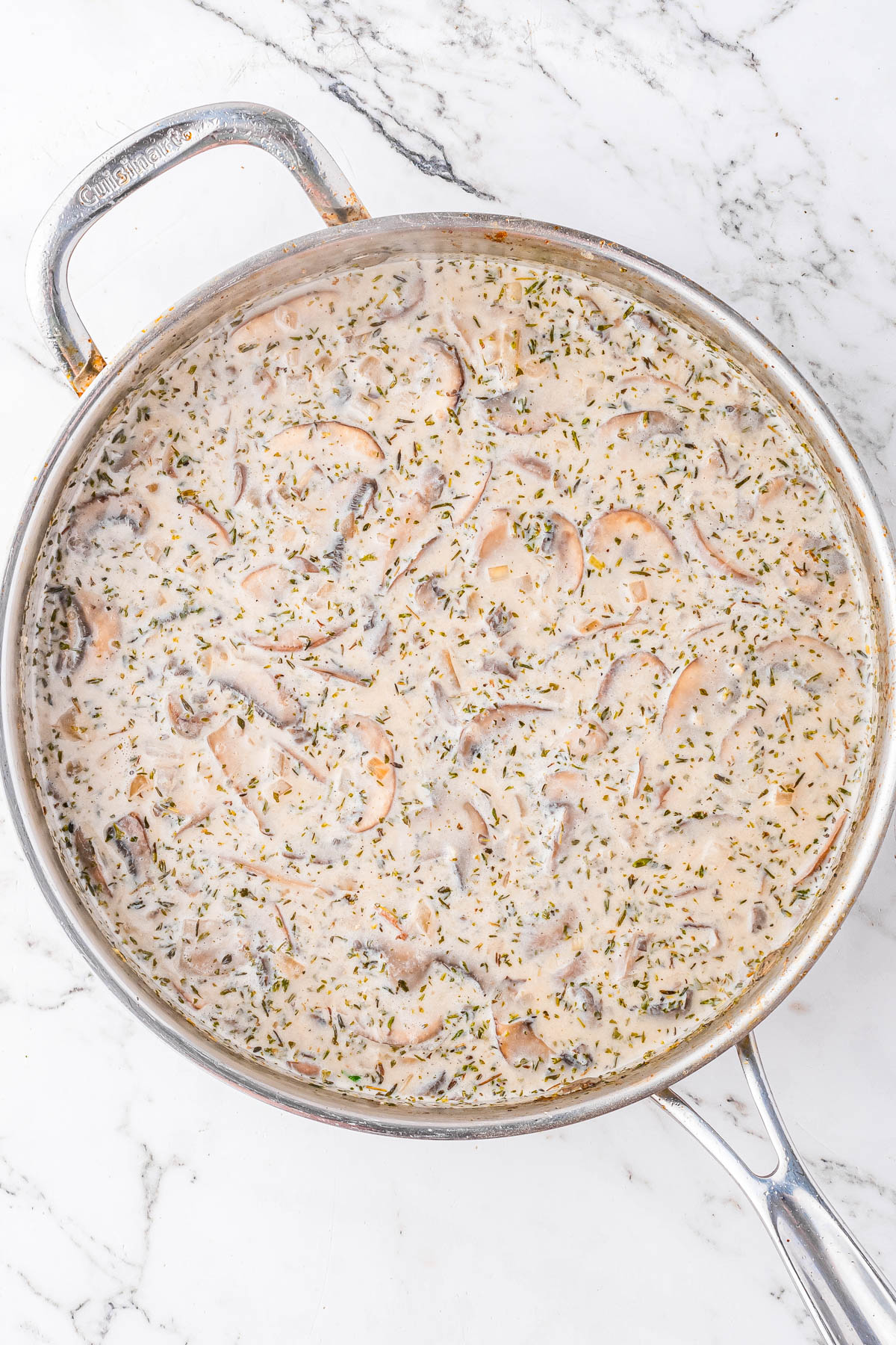 A stainless steel pan on a marble countertop filled with creamy mushroom sauce containing sliced mushrooms and herbs.
