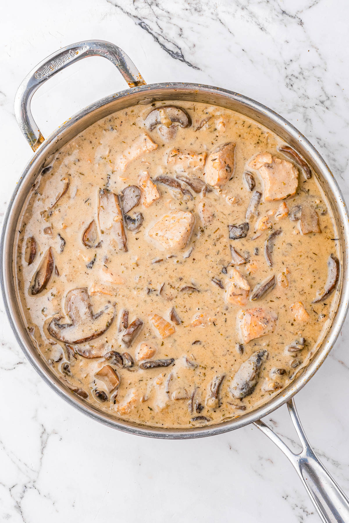 A stainless steel pan filled with creamy mushroom and chicken stew sits on a white marble surface.