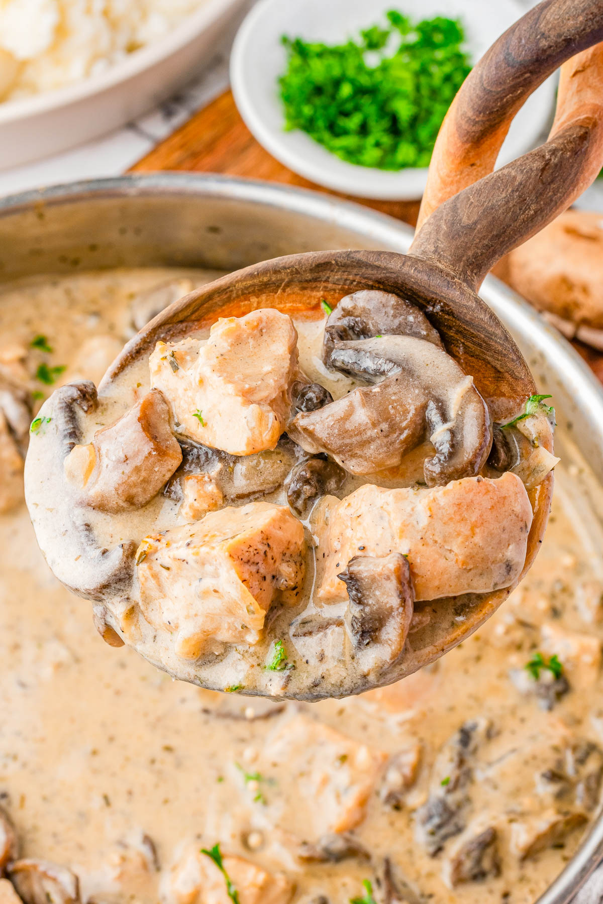 A wooden spoon holds chunks of salmon and mushrooms in a creamy sauce, hovering over a pan filled with the same mixture.