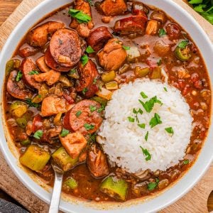 A bowl of gumbo with sausage, chicken, vegetables, and a serving of white rice, garnished with chopped parsley.