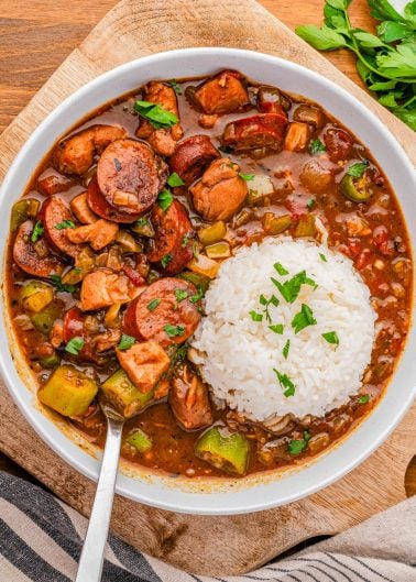 A bowl of gumbo with sausage, chicken, vegetables, and a serving of white rice, garnished with chopped parsley.