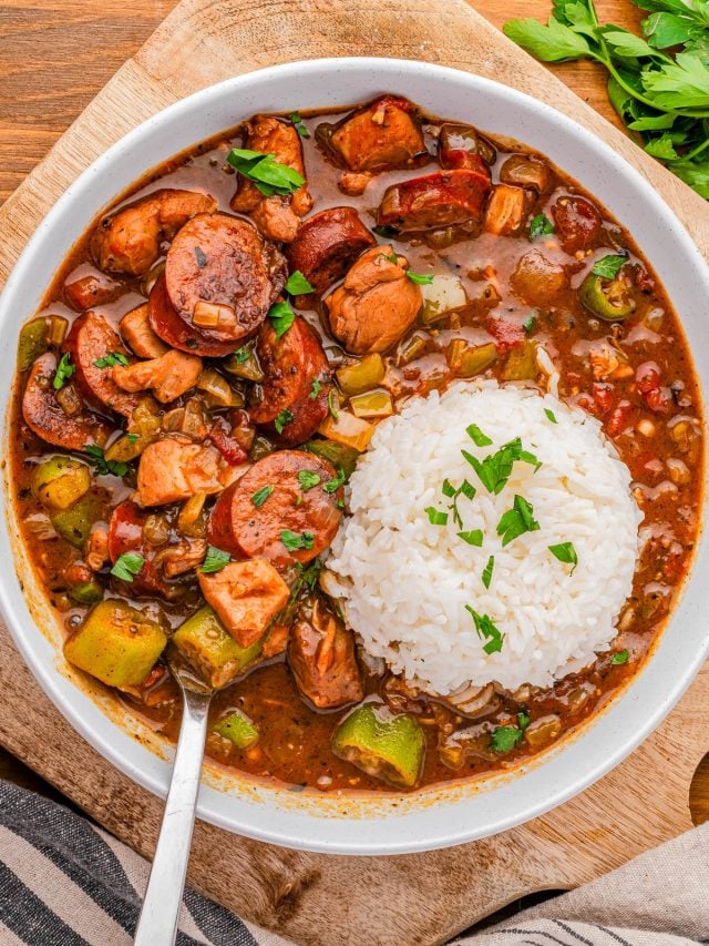 A bowl of gumbo with sausage, chicken, vegetables, and a serving of white rice, garnished with chopped parsley.