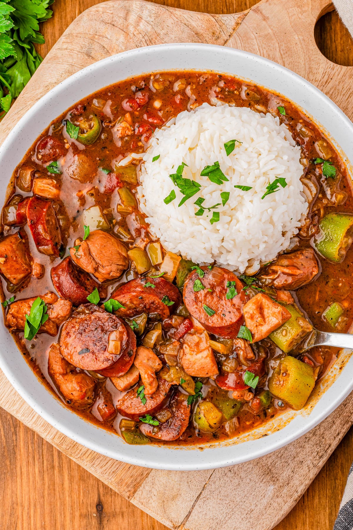 A bowl of gumbo with sausage, chicken, vegetables, and a serving of white rice, garnished with chopped parsley, on a wooden board.