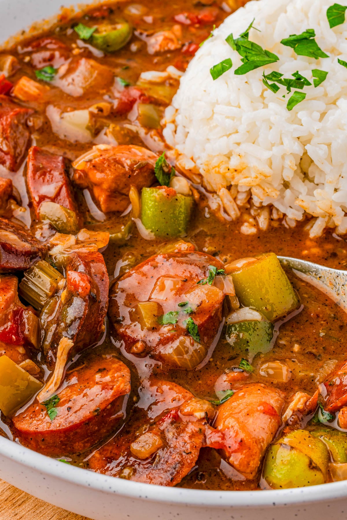 A bowl of gumbo with sliced sausage, vegetables, and shredded meat in a brown sauce, served with a mound of white rice topped with chopped parsley.