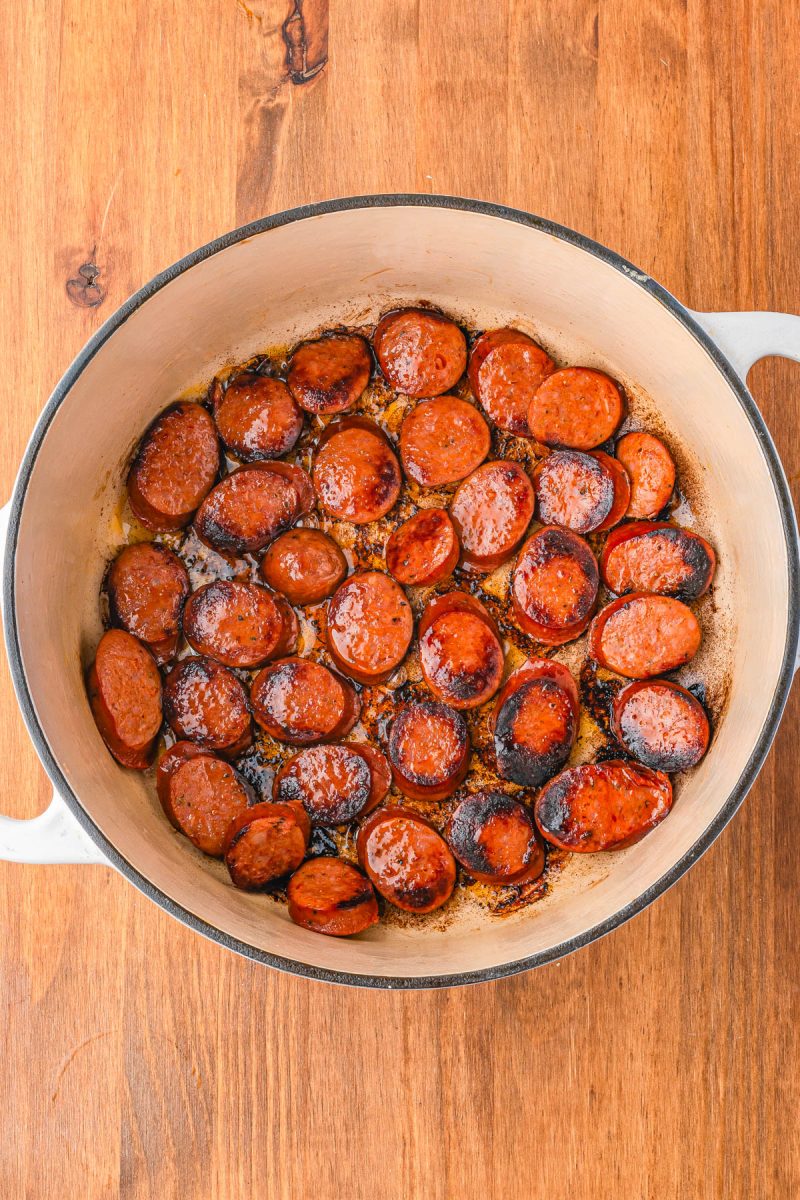A white pot on a wooden surface contains sliced sausage pieces browned and sizzling in oil.