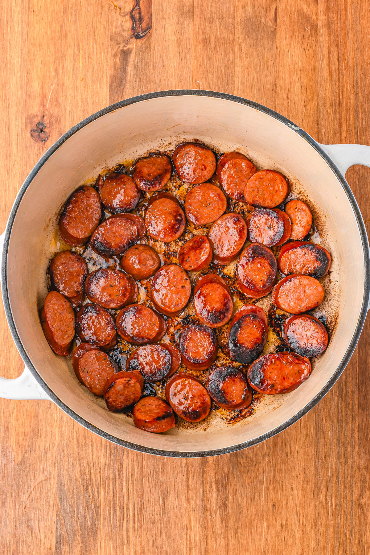 A white pot on a wooden surface contains sliced sausage pieces browned and sizzling in oil.