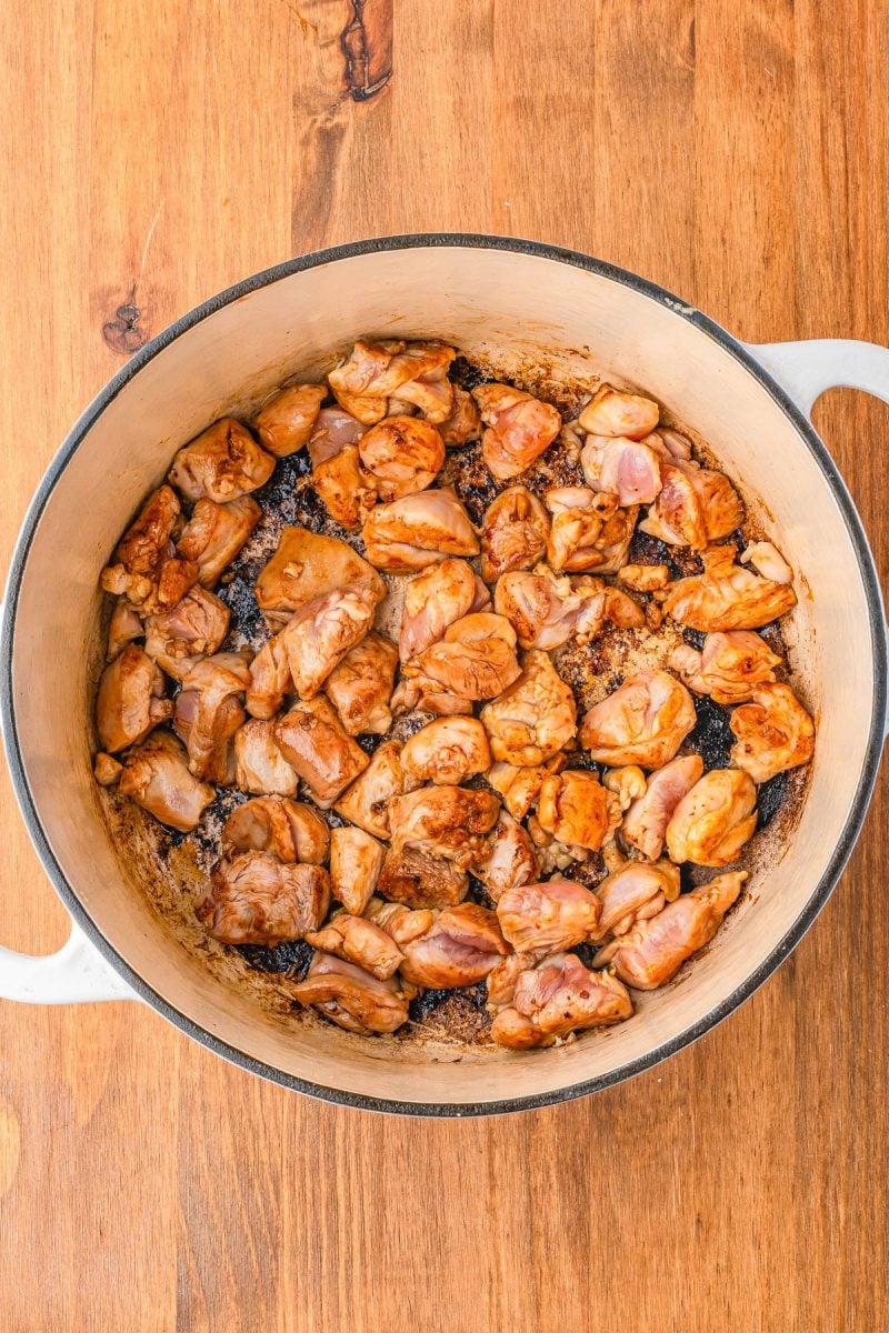 Chunks of seasoned chicken are browning in a large white pot on a wooden surface.