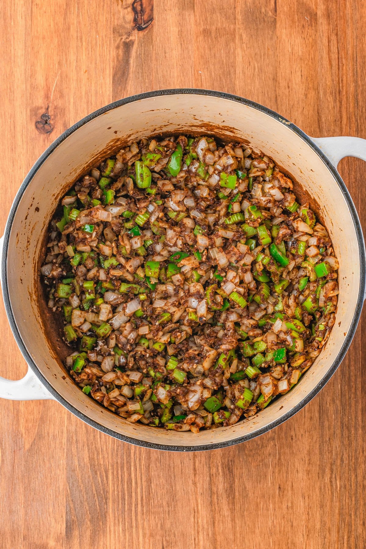 Chopped onions and green bell peppers sautรฉ with ground beef in a large white pot on a wooden surface.