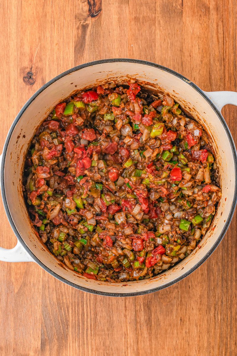 A pot of cooked vegetable mixture with tomatoes, onions, and green peppers on a wooden surface.