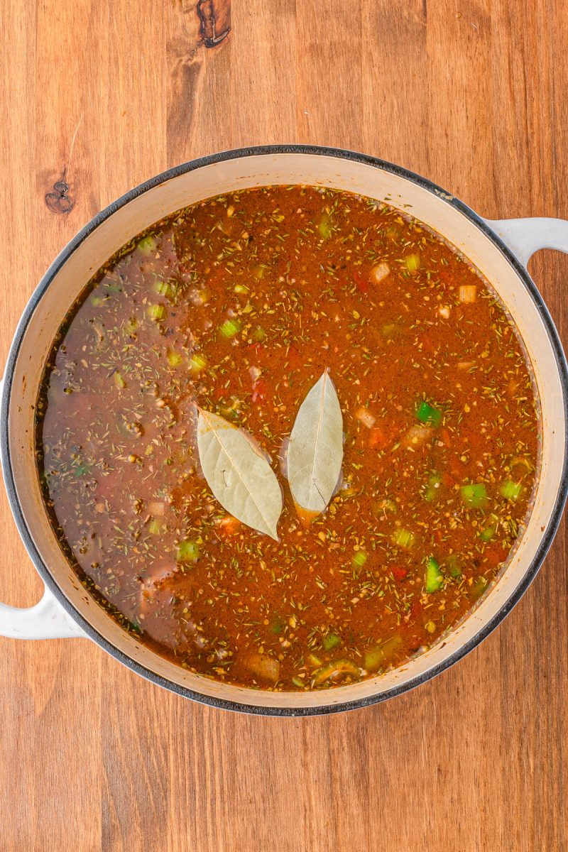 A pot of seasoned broth with chopped vegetables and two bay leaves on top, set on a wooden surface.