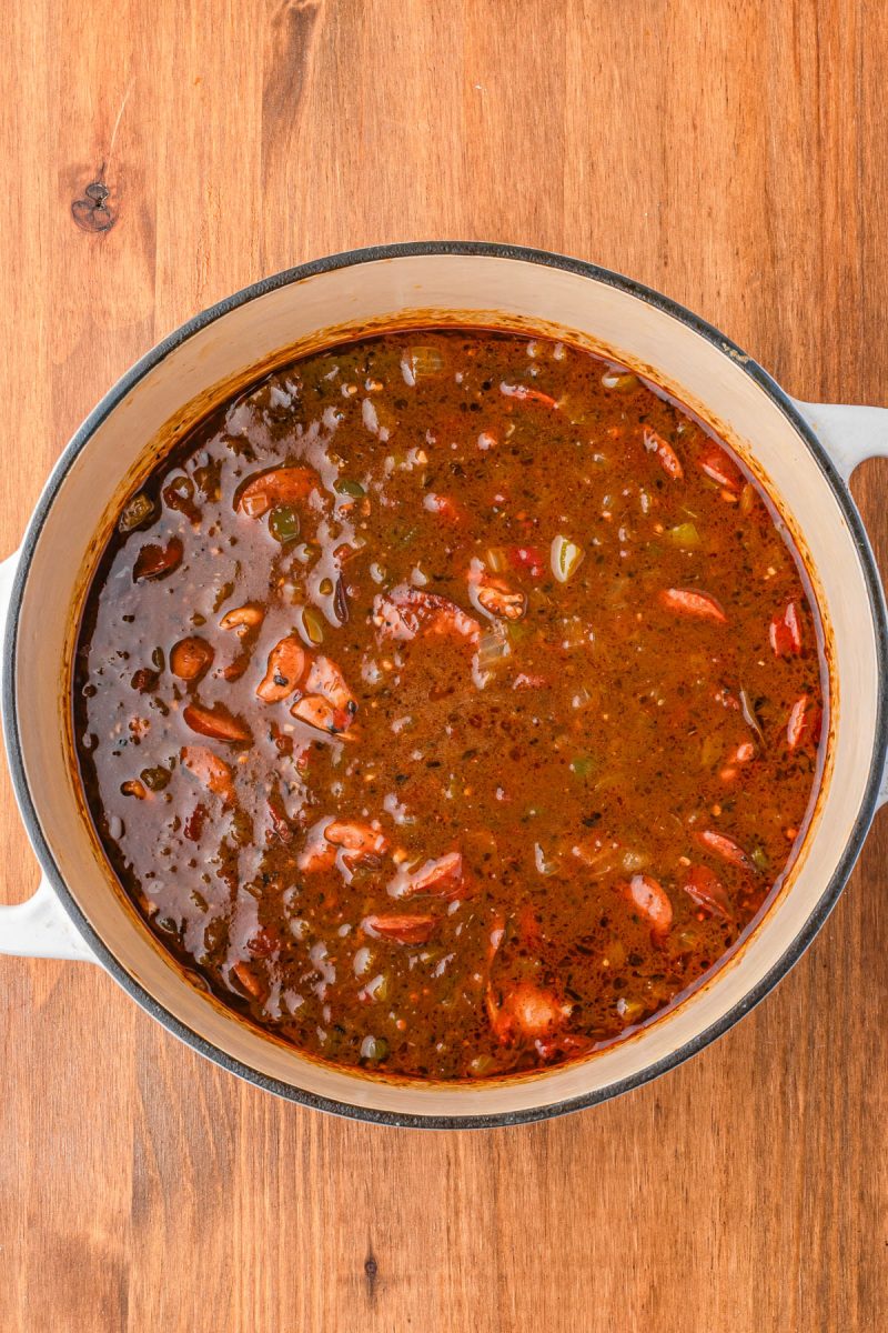 A large pot filled with a dark, chunky stew containing visible pieces of meat, vegetables, and herbs, sits on a wooden surface.