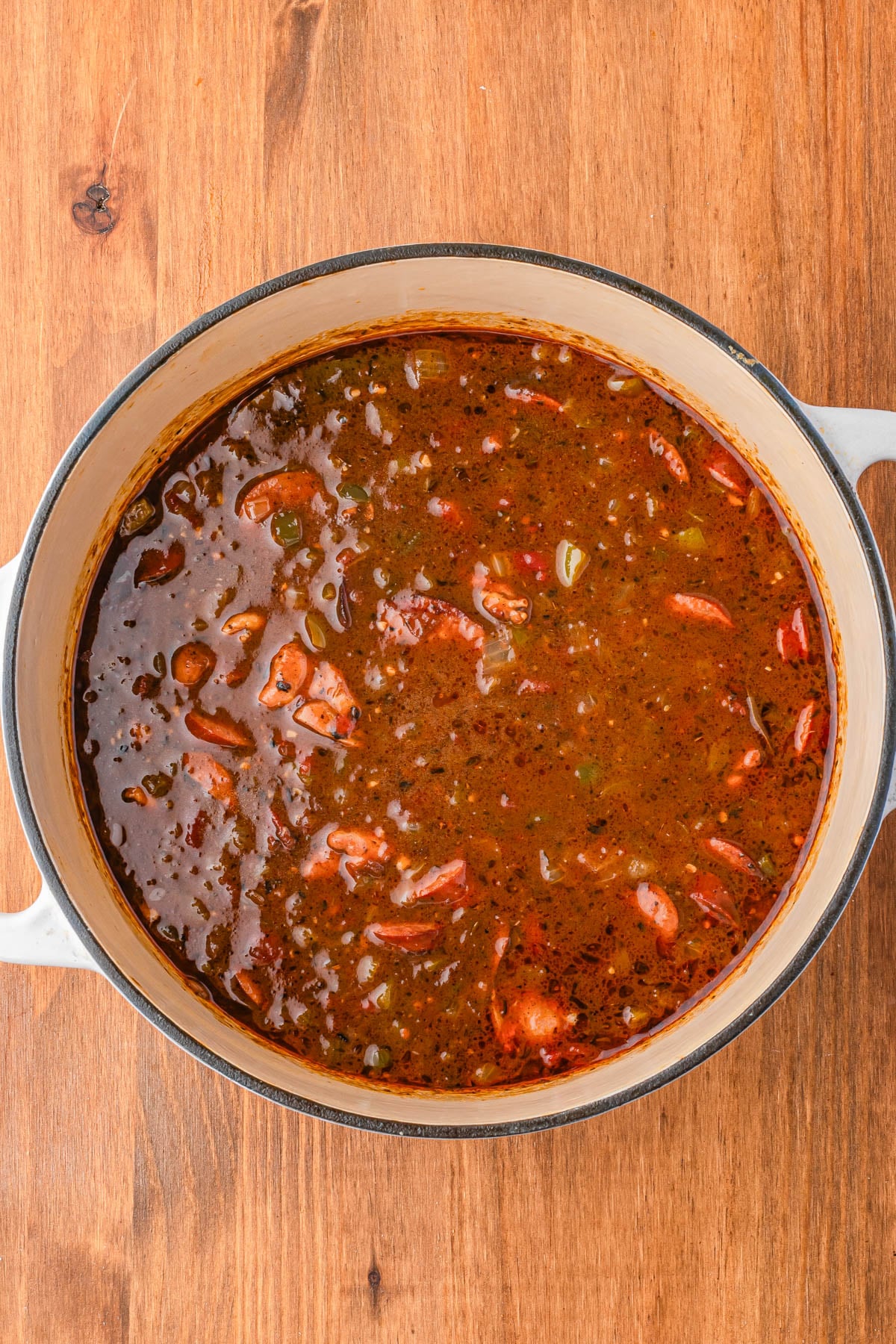 A large pot filled with a dark, chunky stew containing visible pieces of meat, vegetables, and herbs, sits on a wooden surface.
