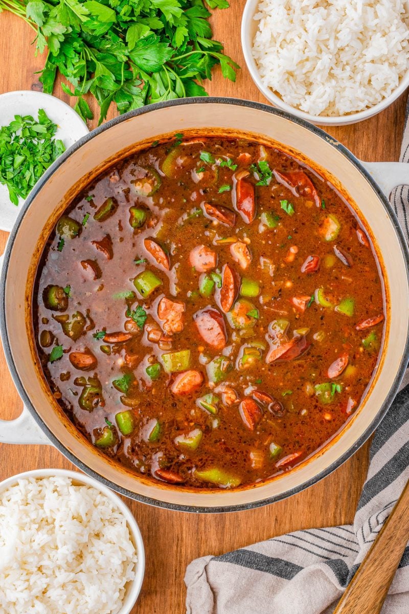 A pot of sausage and vegetable gumbo sits on a wooden surface, surrounded by bowls of white rice and chopped herbs.