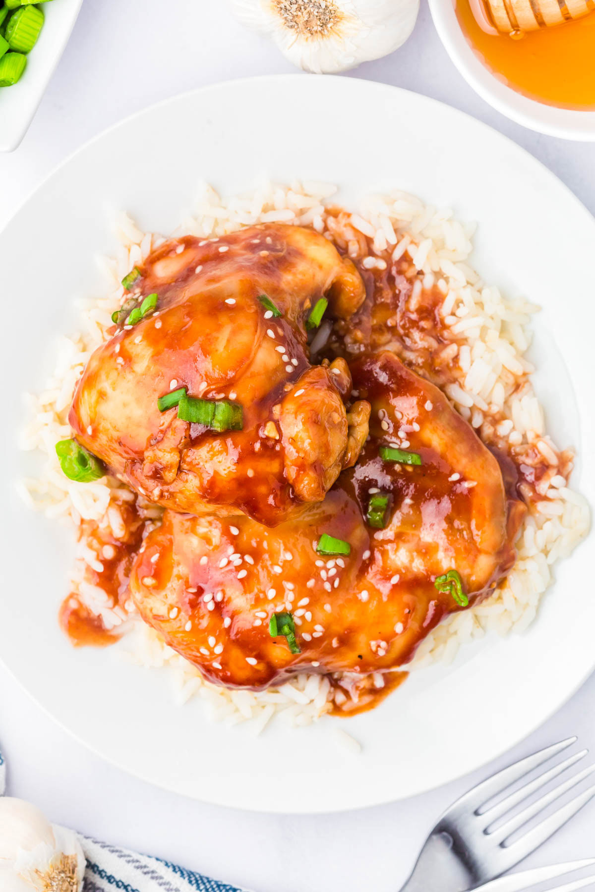 A plate of white rice topped with glazed chicken thighs, garnished with sesame seeds and chopped green onions.
