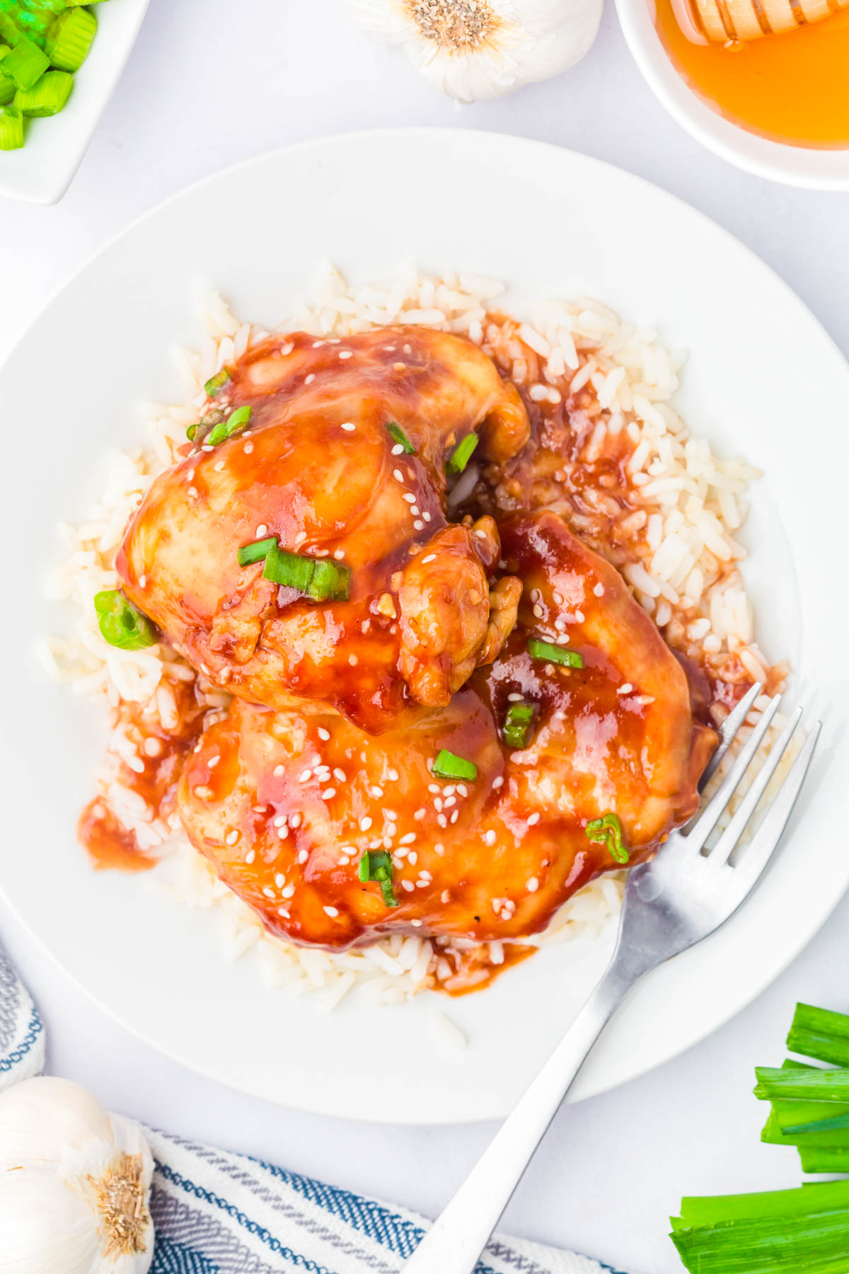 A plate of white rice topped with glazed chicken thighs, garnished with chopped green onions and sesame seeds, with a fork on the side.