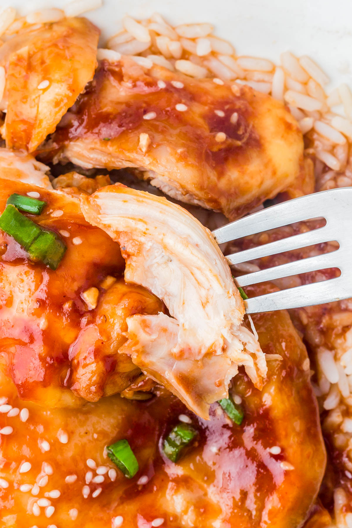 Close-up of a fork pulling a piece of cooked chicken with red sauce, garnished with sesame seeds and chopped green onions, served over rice.