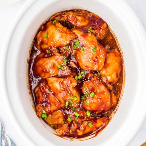 Chicken thighs coated in a glossy brown sauce, garnished with chopped green onions, in a white slow cooker.