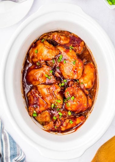 Chicken thighs coated in a glossy brown sauce, garnished with chopped green onions, in a white slow cooker.