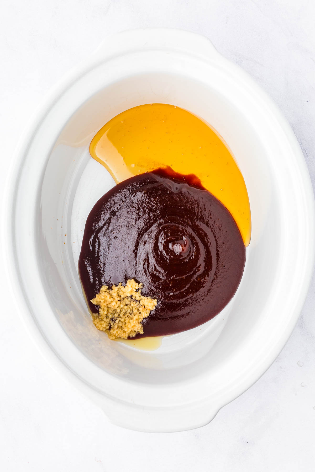 A white slow cooker with honey, barbecue sauce, and minced garlic inside, photographed from above.