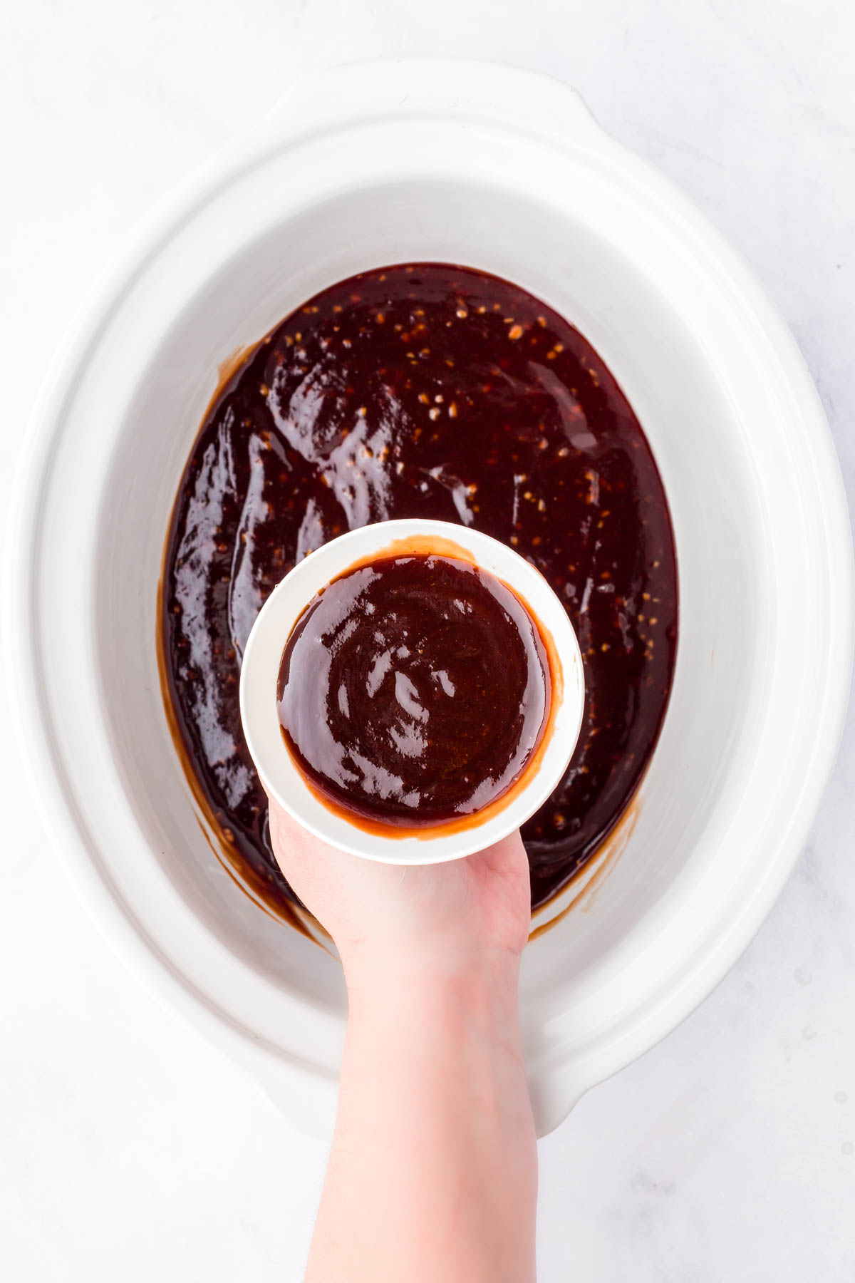 A hand holds a small white bowl of dark barbecue sauce over a slow cooker containing more of the same sauce.