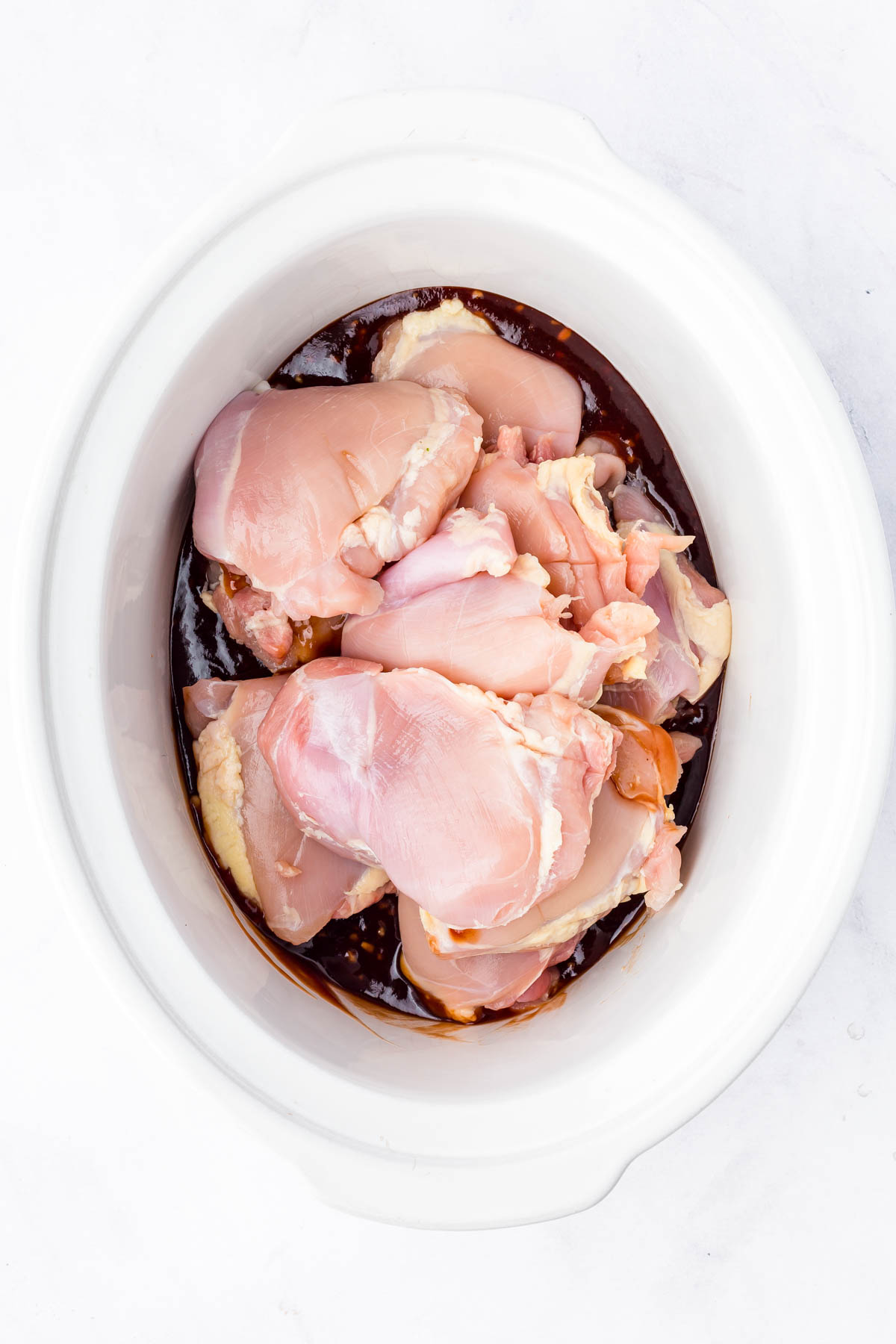 Raw chicken thighs in a white slow cooker with a dark marinade or sauce at the bottom.