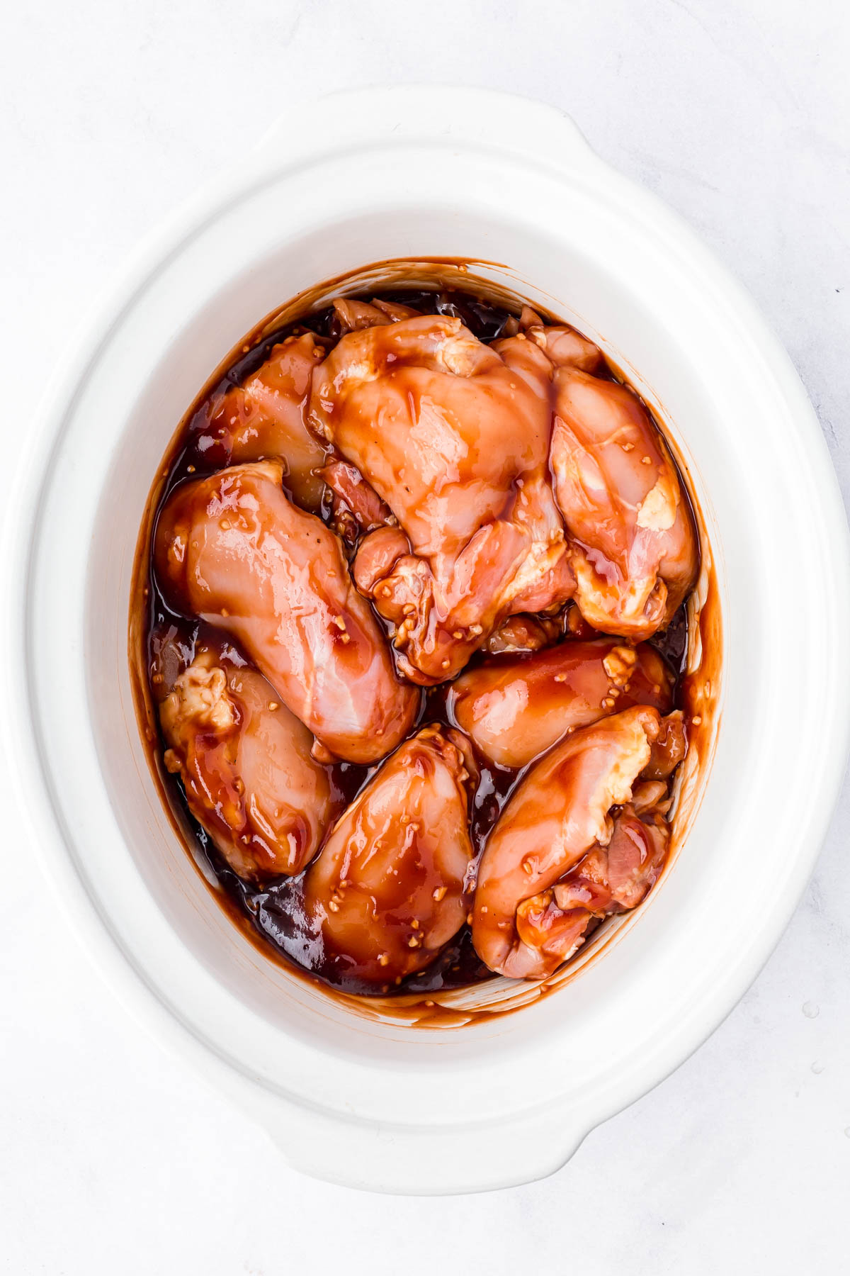 Raw chicken thighs covered in a brown marinade sit in a white slow cooker on a light surface.