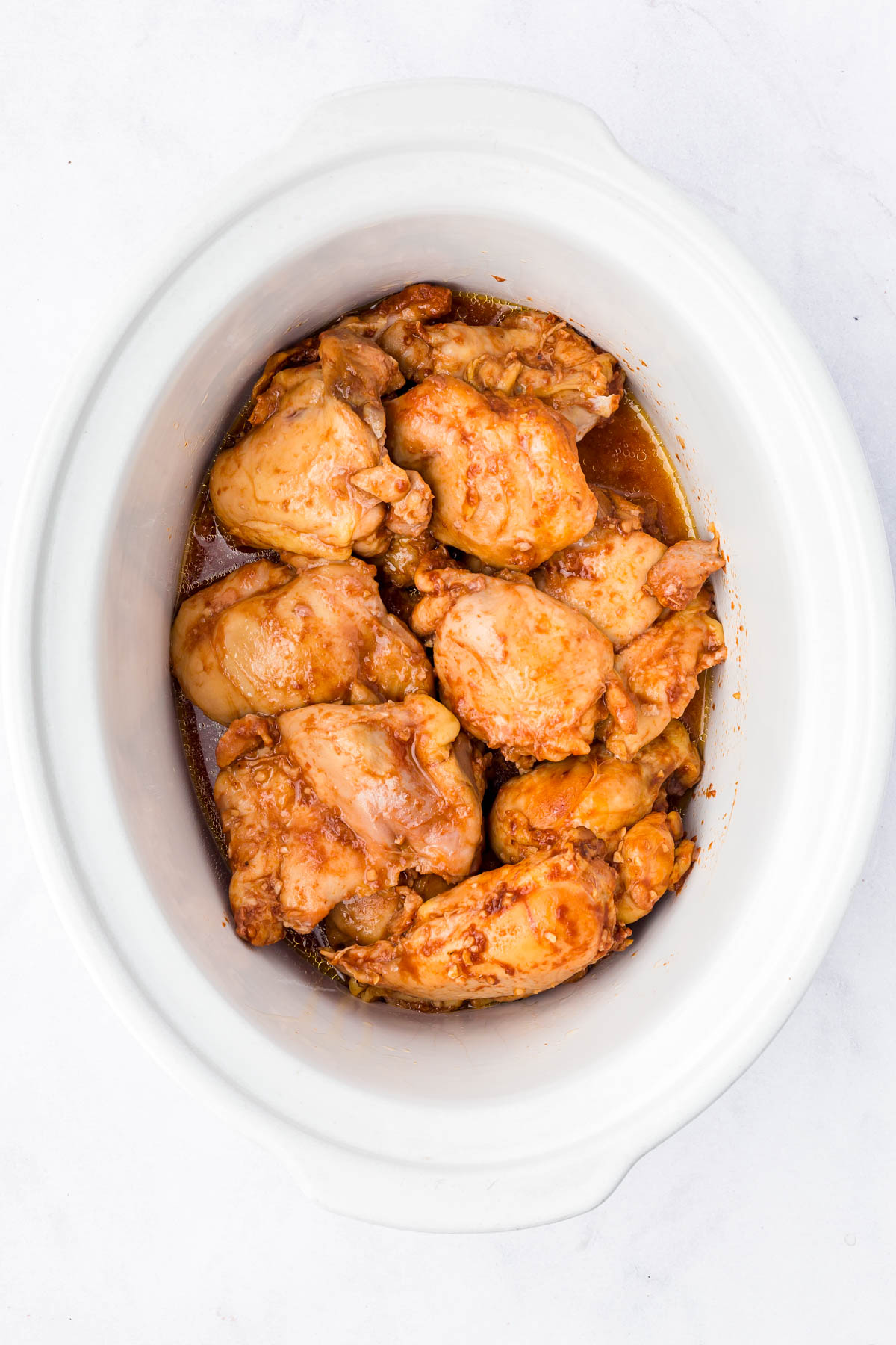 Cooked chicken thighs in sauce arranged inside a white slow cooker on a white surface.