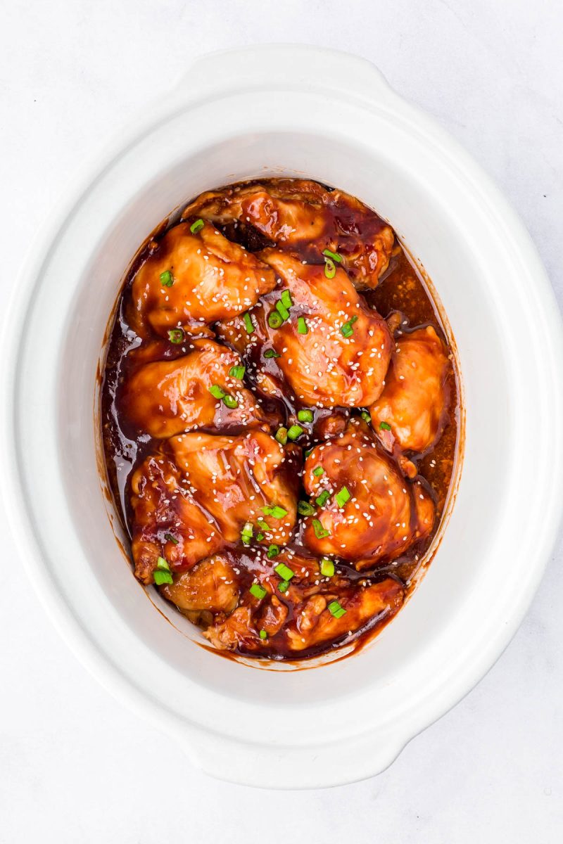 Chicken pieces coated in a thick, glossy sauce topped with chopped green onions and sesame seeds, served in a white oval dish.