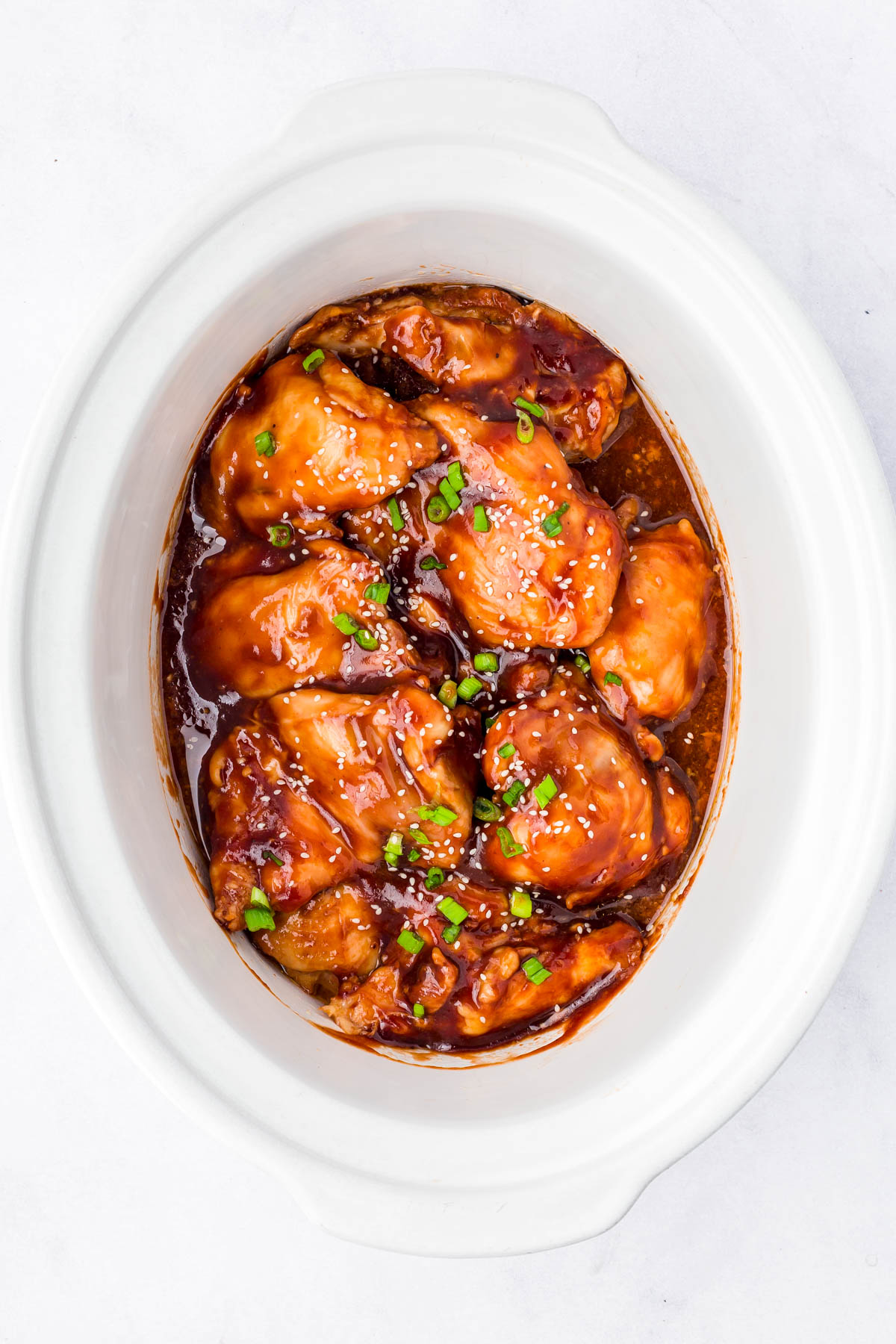 Chicken pieces coated in a thick, glossy sauce topped with chopped green onions and sesame seeds, served in a white oval dish.