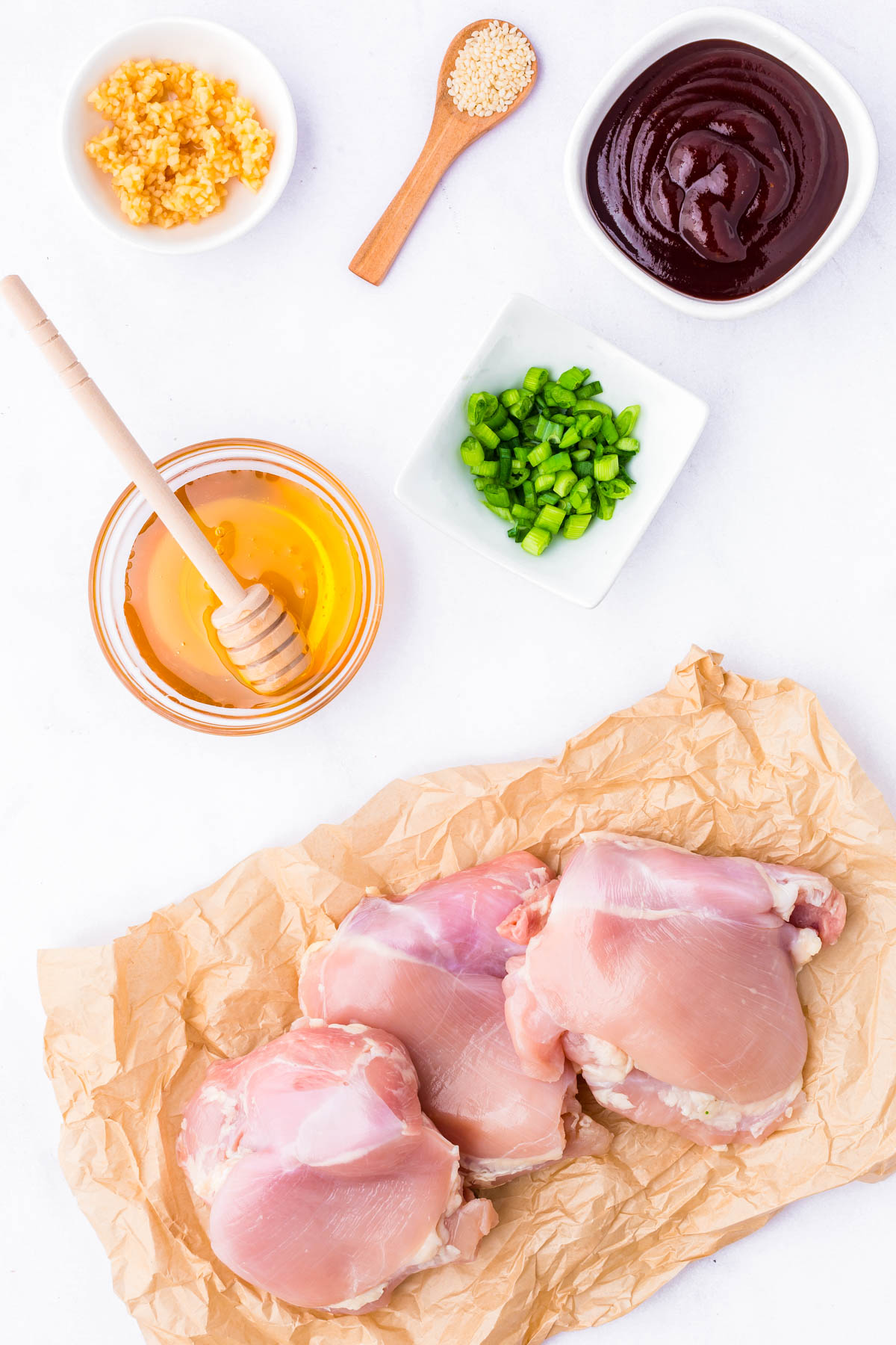 Raw chicken thighs on parchment paper surrounded by bowls of chopped green peppers, minced garlic, honey with a dipper, barbecue sauce, and a spoonful of sesame seeds on a white surface.
