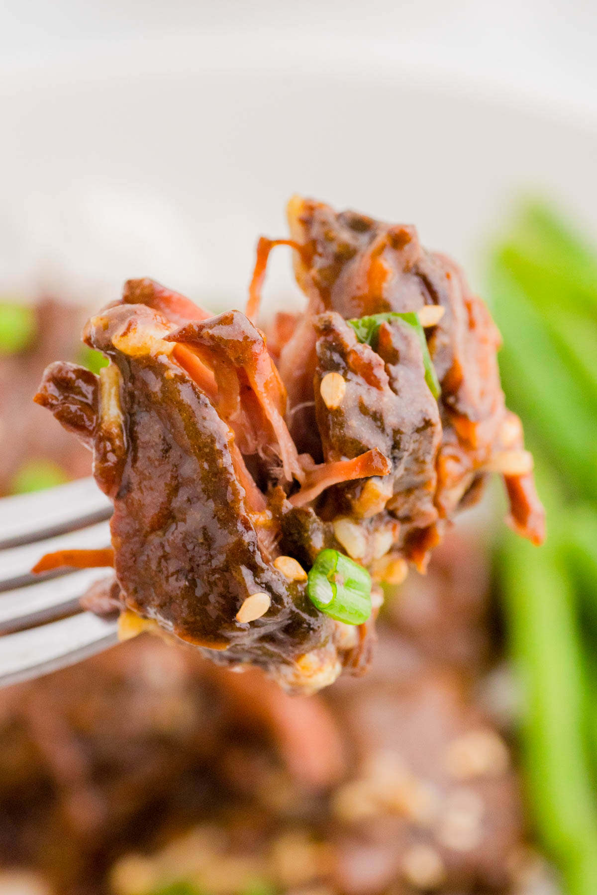 Close-up of a fork holding a bite of saucy beef garnished with sesame seeds and chopped green onions, with green beans blurred in the background.